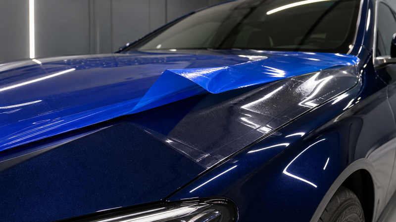 Blue colored cap sheet partially peeled from PPF on a dark-blue performance sedan hood